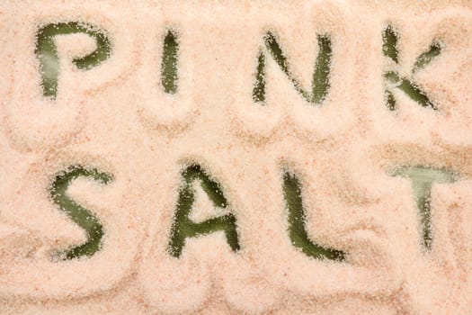 The words PINK SALT cutout from a layer of Himalayan pink salt in a clear glass dish on a green background. The words PINK SALT spelled out in a layer of fine grain Himalayan pink salt close-up.