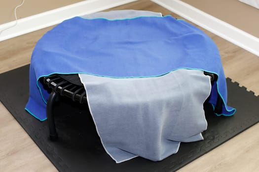 One blue and one gray exercise towel mat covering a round black bungee cord rebounder exercise equipment. A round black rebounder exercise equipment on a black mat covered in exercise towels close-up.
