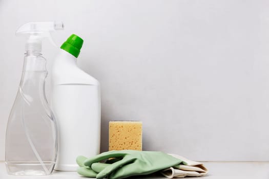 Home cleaning, housekeeping concept. White toilet cleaner bottle, transparent dispenser window cleaner, sanitary ware, washcloth and protective green gloves for housewife on the table, gray background