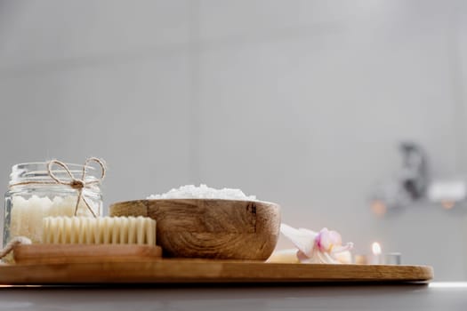 Preparation for hotel spa treatment or home bath procedure. White washbasin in bathroom with accessories on tray. Burning candles, soap, foot brush, glass bottle with sea salt, orchid flower