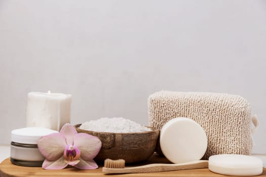 Preparation for hotel spa treatment or home bath procedure. Gray bathroom with accessories on tray on washbasin. Burning candle, washcloth, soap, foot and teeth brush, cream bottle, sea salt in bowl