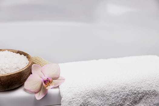 Preparation for hotel spa treatment, home bath procedure. White washbasin in bathroom, accessories on tray. Foot or body brush, white towel, sea salt in bamboo bowl, orchid flower. High quality photo