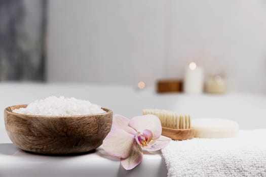 Preparation for hotel spa treatment, home bath procedure. White washbasin in bathroom, accessories on tray. Burning candles, soap, foot brush, towel, glass bottle with sea salt, orchid flower