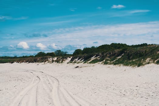 Dunes, overgrown with grass in places, car tracks on the sand. Blue sky with clouds.