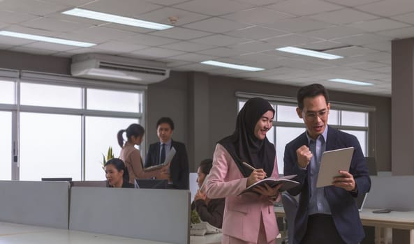 Happy businessman in dark blue suit using digital tablet working with Muslim women wearing hijab making notes while standing in open space office. Business team workplace concept
