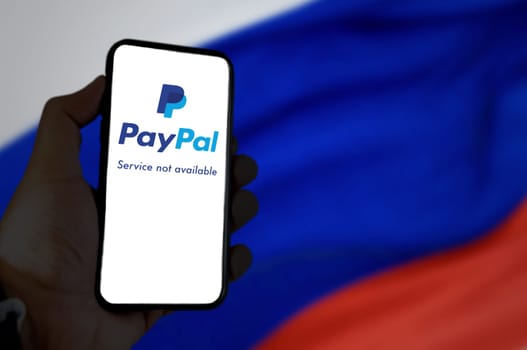 Moscow, RUS, March 2022: Hand holding a mobile phone with the Paypal app on the screen with the Russian flag blurred in the background. In March 2022 Paypal has suspended all its services in Russia