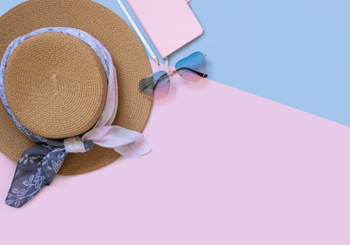 Straw hat, notebook, pensil and heart sunglasses top view on light pink and blue. Female summer clothes and accessories flat lay. Hardcover textbook mockup. Girly travel concept