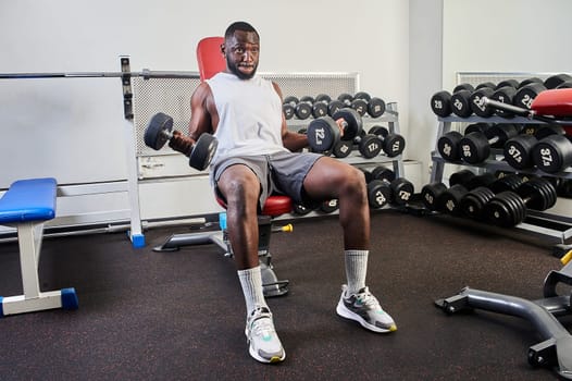 An African American man shakes his biceps while sitting on a bench in the gym. High quality photo