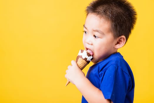 Happy portrait Asian child or kid cute little boy attractive laugh smile playing holds and eating sweet chocolate ice cream waffle cone, studio shot isolated on yellow background, summer concept