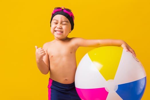 Summer vacation concept, Portrait Asian happy cute little child boy smiling in swimsuit hold beach ball and showing finger thumb up for good sign, studio shot isolated yellow background