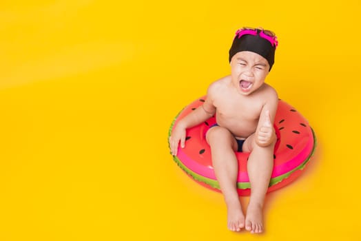 Summer vacation concept, Portrait Asian happy cute little child boy wear goggles, swimsuit hold beach watermelon inflatable ring, Kid have fun sit in inflatable, studio shot isolated yellow background