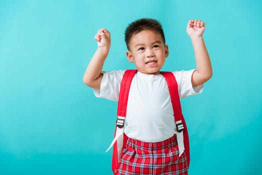 Back to school. Portrait happy Asian cute little child boy in uniform smile raise hands up glad when go back to school, isolated blue background. Kid from preschool kindergarten with school backpack