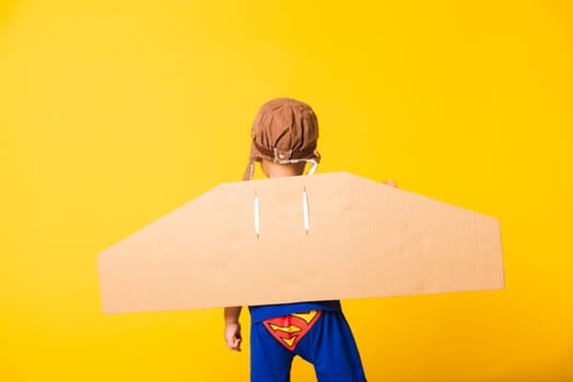 Back of Happy Asian handsome funny child or kid little boy smile wear pilot hat play raise hand up with toy cardboard airplane wings flying, studio shot isolated yellow background, Startup freedom