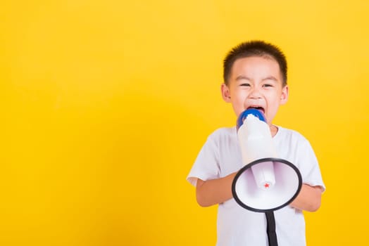 Asian Thai happy portrait cute little cheerful child boy holding and shouting or screaming through the megaphone her looking to camera, studio shot isolated on yellow background with copy space