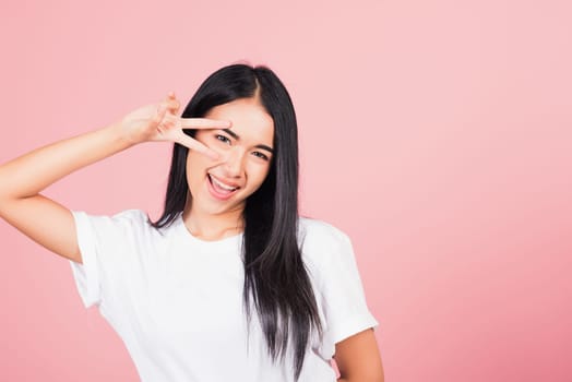 Asian happy portrait beautiful cute young woman teen smile standing showing finger making v-sign victory symbol near eye looking to camera studio shot isolated on pink background with copy space