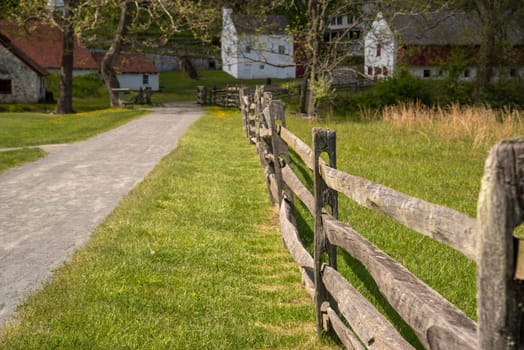 White defocused buildings and barns are located in the background at the end of a walking path surrounded by green fields and a brown split rail fence. Shot in natural sunlight with no people and copy space.