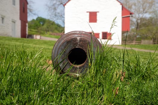 A cannon in the grass at a colonial American iron forge and surrounding village. Closeup look down the barrel, whitewashed buildings in the background of this historic site. Copy space, no people, shot in natural light