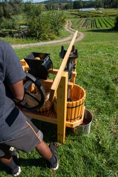 A young African-American man operates a wooden cider press at the top aof a grassy hill with a dirt road and family farm in the background