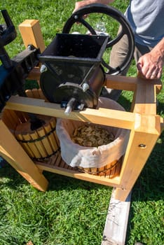 A caucasian man turns the fruit grinder flywheel of an outdoor wooden apple cider press on a sunny day. Shot in natural light with copy space.