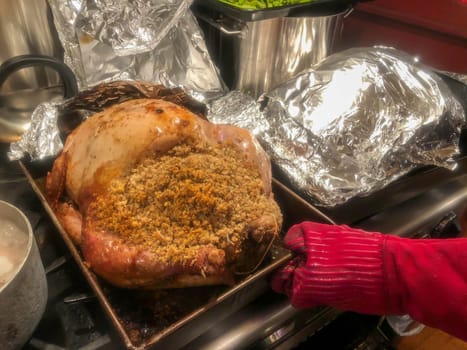 Foil is removed from turkey for browning. Second covered turkey on right, red hot mitt holds the roasting pan with a gorgeous stuffed turkey inside and silver pots on kitchen stovetop.
