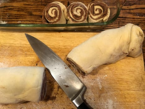 Beautiful hand rolled cinnamon buns with knife and cut rolls in baking dish. High angle view of rustic home bakery natural look with quality steel knife blade, glass dish, and wooden cutting board