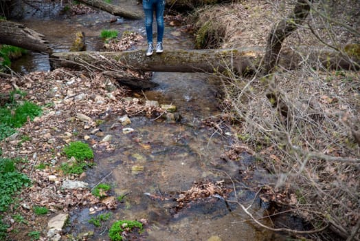 A person in athletic shoes stands on a fallen tree footbridge over a woodland stream in an early spring landscape. Bright new green growth on creek banks and clean flowing water. Natural light, with copy space.