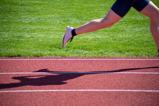 Motion blur on runner's foot and leg as it moves across the image with the runner's shadow on the track. Crisp horizontal lane lines and green grass background in natural sunlight with copy space and no people.