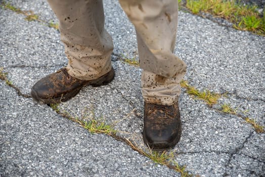 A man's feet in work boots and pants splattered with dirt and grease stands on a proken asphalt road. Natural light with copy space.