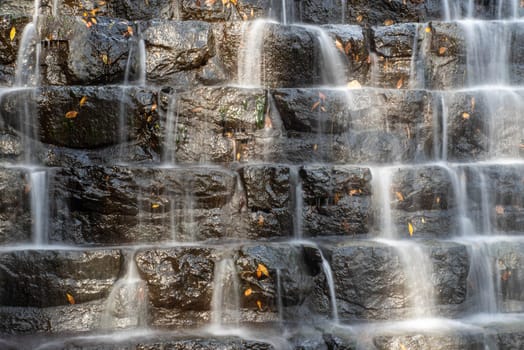 Autumn leaves cling to the black rocks of a tiered man made waterfall. Water splashes dynamically and the stones shine in the dappled sunlight. A great nature background.