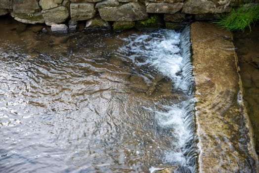 Stone lined man made channel of flowing water cascades over an old colonial American Pennsylvania dam with texture water ripples and copy space in natural light with no people.