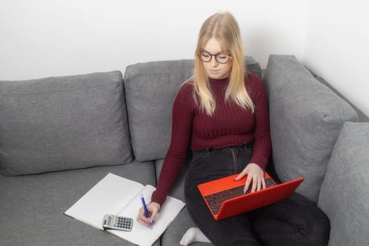 Young woman in glasses sit on couch and work on laptop writing on paper documents, pay bills or taxes, use computer at home