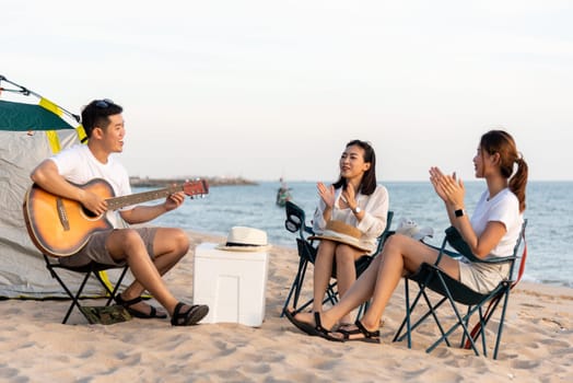 Happy friend have fun playing guitar and clap in camp they smiling together in holiday on sand beach near camping tent vacation time at sunset, Young Asian group woman and man in summer travel outdoor