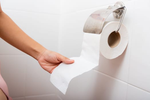 Closeup hand pulling toilet paper roll in holder for wipe, woman sitting on toilet she taking and tearing white tissue on wall to towel clean in bathroom, Healthcare concept