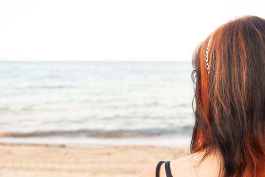 Young woman with red and black long hair standing at the beach, looking at the sea, rear view, horizontal
