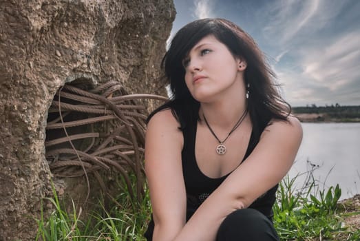 Young gothic emo punk girl sitting at the lake, black hair, wearing black clothes, looking up, horizontal