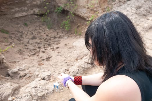Young alternative emo girl with black hair and black clothes, sitting outdoors on the ground during daylight, looking down, side view, horizontal