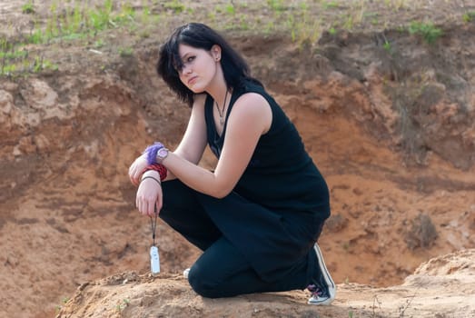 Punk emo girl, young adult with long black hair, kneeling on a hill outdoors in the nature, holding a camera in her left hand, looking at camera, horizontal
