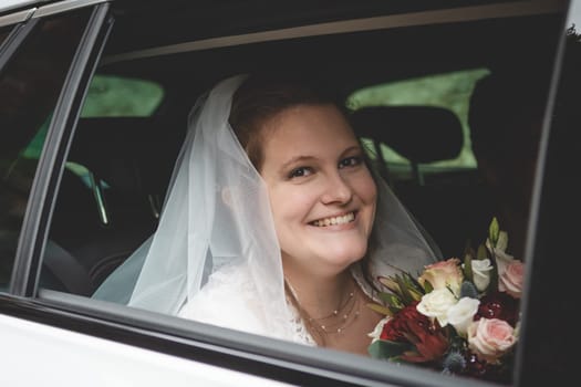 Bride with brown hair, sitting in car on her way to her wedding, holding flowers, smiling and looking at camera through a car window