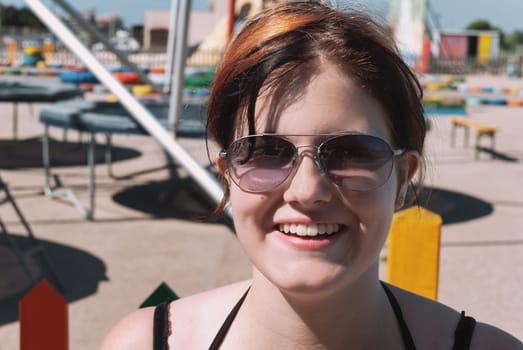 Young woman with black orange hair, wearing sunglasses, smiling, looking at camera while standing in front of a trampoline park in majorca, close-up