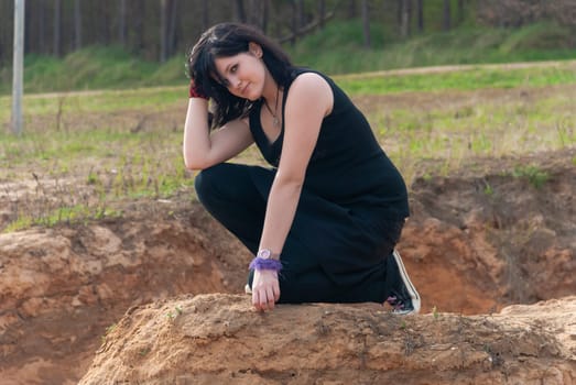 Punk emo girl, young adult with long black hair, kneeling on a hill outdoors in the nature, looking at camera, horizontal