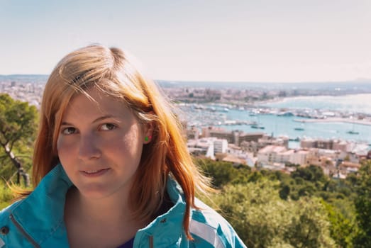 Young woman with blond orange colored hair looking at camera while standing on a observation point with palma, majorca in the background, horizontal