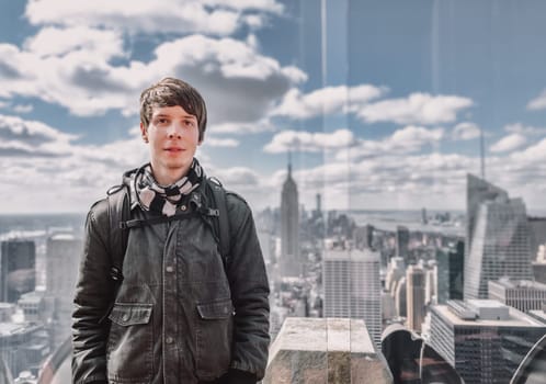 Young adult man with brown hair and beard looking at camera while standing on a observation tower in front of new york city skyline, usa, america