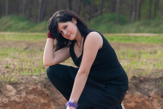 Punk emo girl, young adult with long black hair, kneeling on a hill outdoors in the nature, looking at camera, horizontal