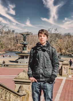 Young white adult man with brown hair standing in front of a fountain on stairs at central park new york, wearing a jacket and scarf, winter in new york city, usa, vertical