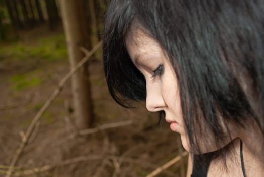 Punk emo girl, young adult with black hair and eyeliner, looking down, forest, close-up, horizontal