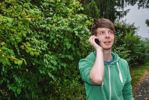 Young man with brown hair and a beard is talking on his smartphone in the forest, wearing a green sweater