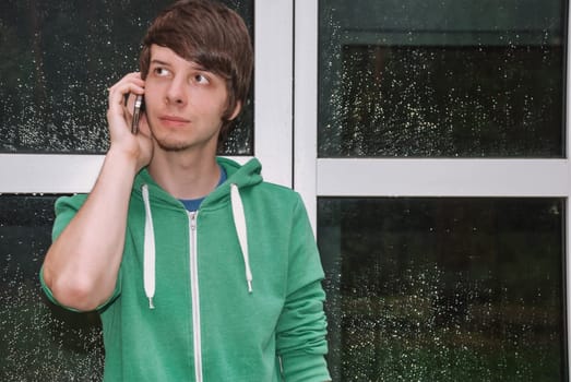young man with brown hair and a beard is talking on the phone with his smartphone in front of his door with raindrops, wearing a green sweater