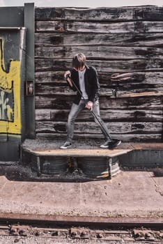 Young adult singer, with brown long hair and beard performing, rock musician with microphone outdoors in front of a wooden wall, standing, looking away, wearing jeans and t-shirt