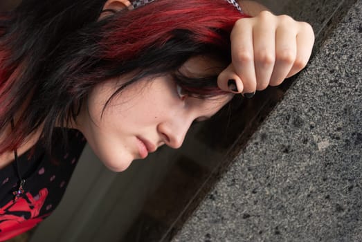 Punk emo girl, young adult with black red hair, holindg her head against a concrete wall, close-up, horizontal