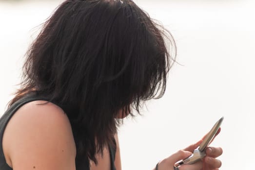 Punk emo girl with long black hair and eyeliner, young adult using her mobile phone while sitting outdoors at the lake, holding a camera in her right hand, horizontal